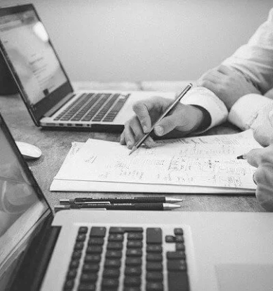 Dois notebooks e um homem com um lapis escrevendo em um papel no meio dos dois notebooks.